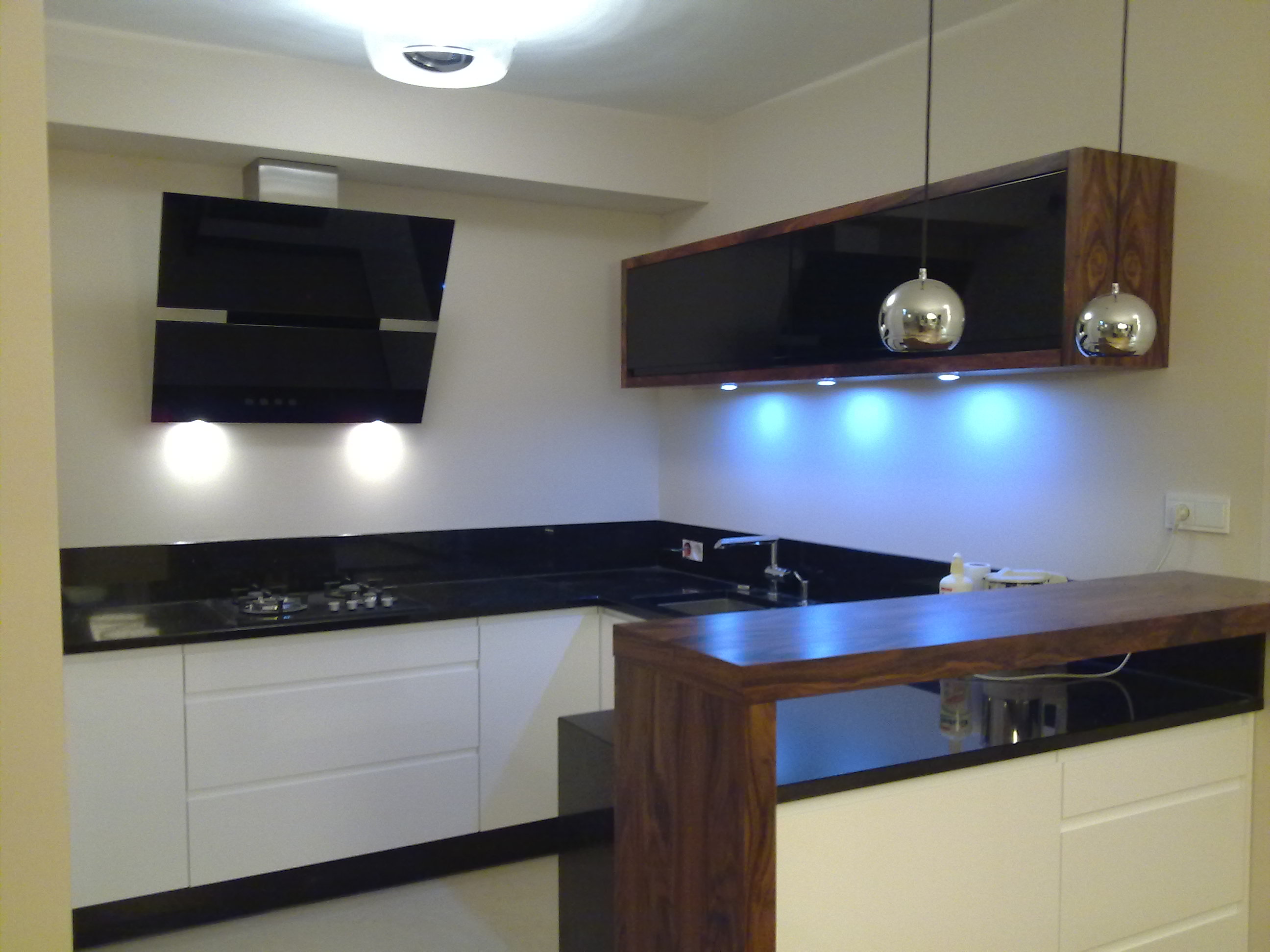 Nowoczesna kuchnia na wymiar z białymi frontami, czarnym blatem i drewnianym barkiem, oświetlona lampami wiszącymi i podświetleniem szafek.